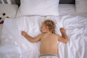 Tvååring sover på mage i en vit säng under dagen